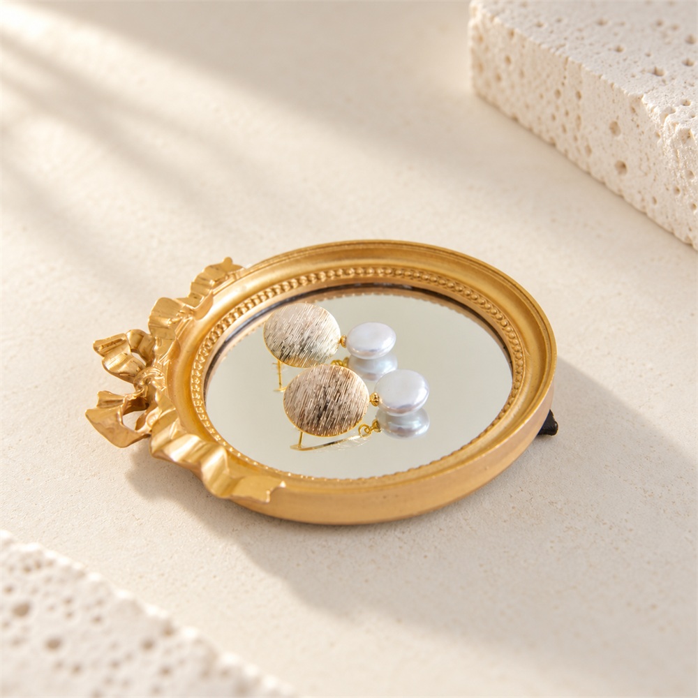 Brushed gold-tone disc and button pearl earrings reflected in a round mirror