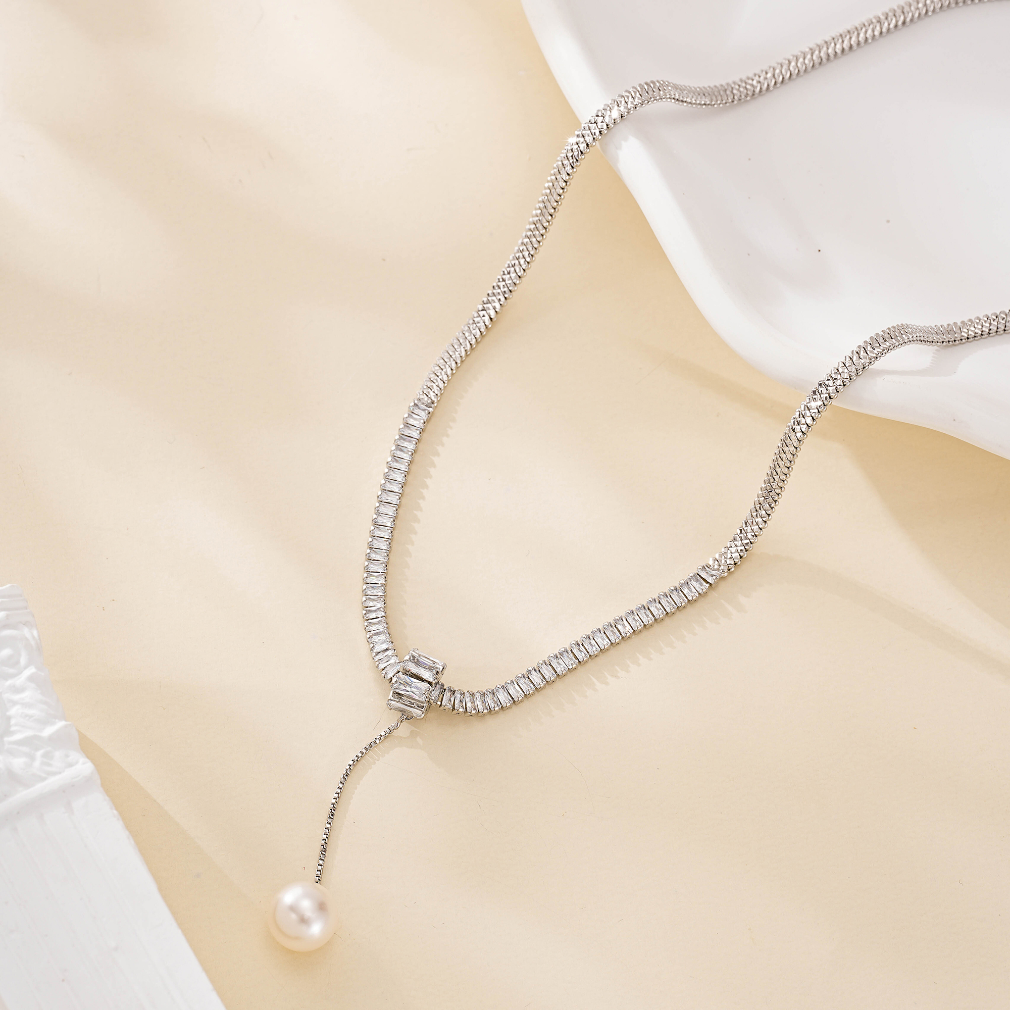 Silver-tone snake chain necklace with a long pearl drop draped beside a white dish